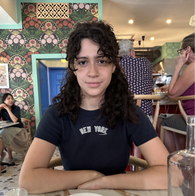 A photograph of Aliana, a Latina girl with curly hair sitting at a table with her arms crossed in front of her, looking at the camera.